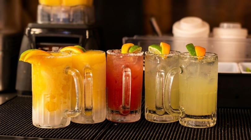 Four colorful margaritas, two yellow, one red, and one light green, garnished with citrus slices.