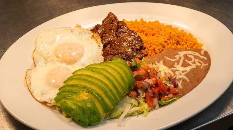 A white plate with steak, two fried eggs, rice, refried beans, sliced avocado, and pico de gallo.