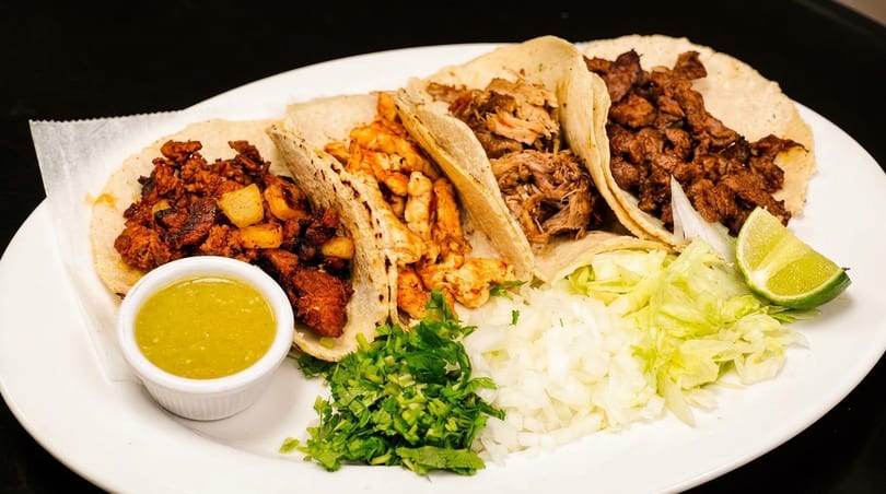 A plate of four diverse tacos with various fillings, salsa, cilantro, onions, lettuce, and lime.